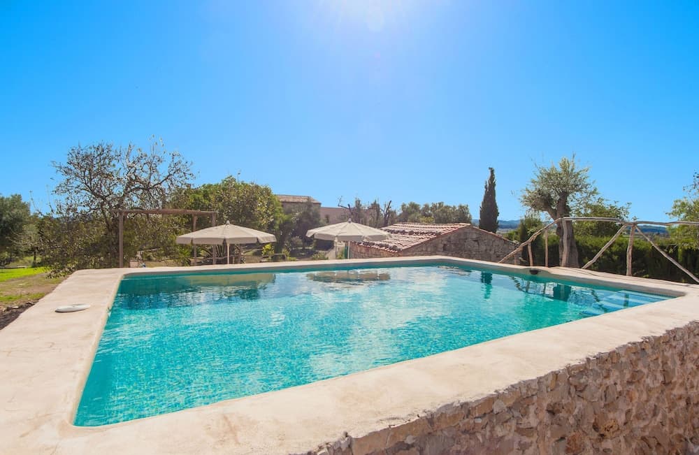 A serene countryside villa features a sparkling pool surrounded by lush greenery under a bright blue sky, ideal for a relaxing vacation retreat.