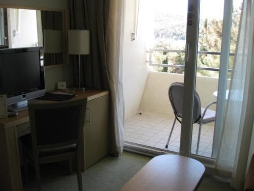 A cozy hotel room with a desk, chair, and television opens to a serene balcony overlooking lush greenery, ideal for travelers seeking relaxation and comfort.
