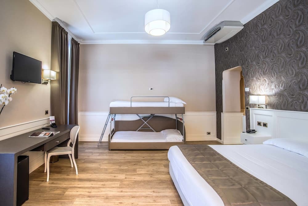 A cozy hotel room features a bunk bed, a double bed, a desk with a chair, and a wall-mounted TV, creating a comfortable and functional space ideal for family travelers seeking convenience and comfort.