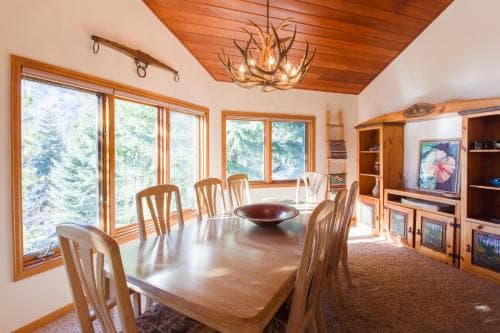 A cozy cabin dining area bathed in natural light from large windows overlooking a forest, featuring rustic wooden furniture, an antler chandelier, and warm decor, ideal for a serene getaway retreat.