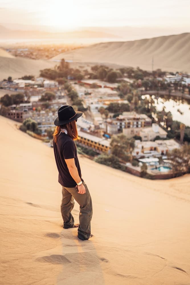 A traveler in a black hat stands atop a sand dune overlooking a serene oasis town at sunset, capturing the tranquil beauty of desert exploration.