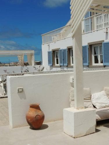 A serene rooftop terrace features white walls, blue shutters, a cozy lounge area, and an ancient clay pot, perfect for a tranquil Mediterranean getaway.