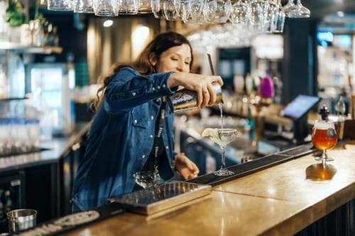 A bartender pours a cocktail into a martini glass at a cozy bar, surrounded by elegant glassware and warm lighting, ideal for travelers seeking a sophisticated nightlife experience.