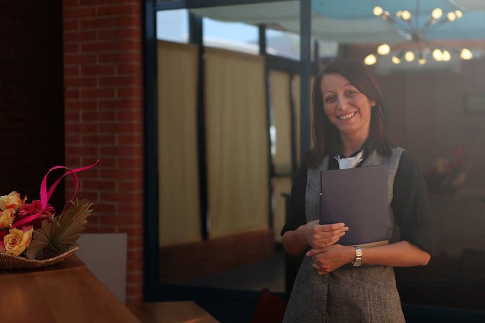 A warmly lit café entrance features a smiling hostess holding a menu near a vibrant floral arrangement, inviting travelers to explore cozy dining experiences.