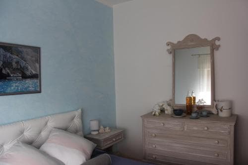 A cozy bedroom with soft blue walls, a vintage dresser adorned with decorative items and a mirror reflecting natural light, creates a serene atmosphere ideal for travelers seeking relaxation.