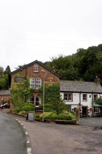 A quaint village scene featuring the Fox & Pheasant Inn, surrounded by lush greenery and charming architecture, evokes a serene and inviting atmosphere perfect for travelers seeking a peaceful countryside retreat.