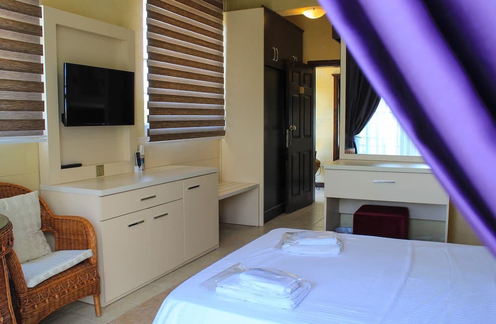 A cozy hotel room features a neatly made bed, wicker chair, flat-screen TV, and wooden blinds, creating a serene atmosphere ideal for travelers seeking relaxation.