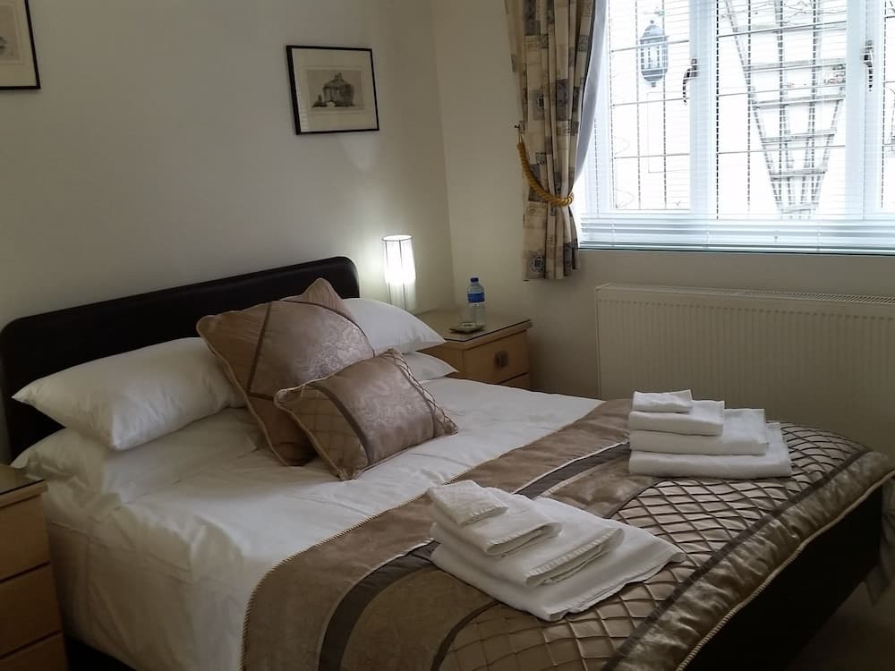 A cozy hotel room features a neatly made bed with white linens and folded towels, a bedside lamp, framed artwork, and a window with blinds, ideal for travelers seeking comfort and relaxation.