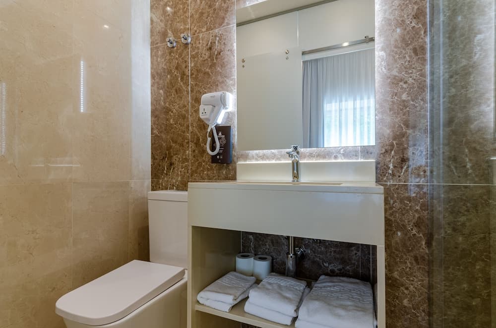 A modern hotel bathroom features sleek marble walls, a wall-mounted hairdryer, neatly folded towels under the sink, and a bright, clean atmosphere ideal for travelers seeking comfort and convenience.