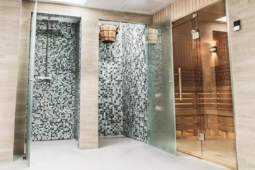 A serene wellness center features a modern sauna room with wooden benches, glass-enclosed showers with mosaic tiles, and ambient lighting, ideal for relaxation and rejuvenation during a spa retreat.