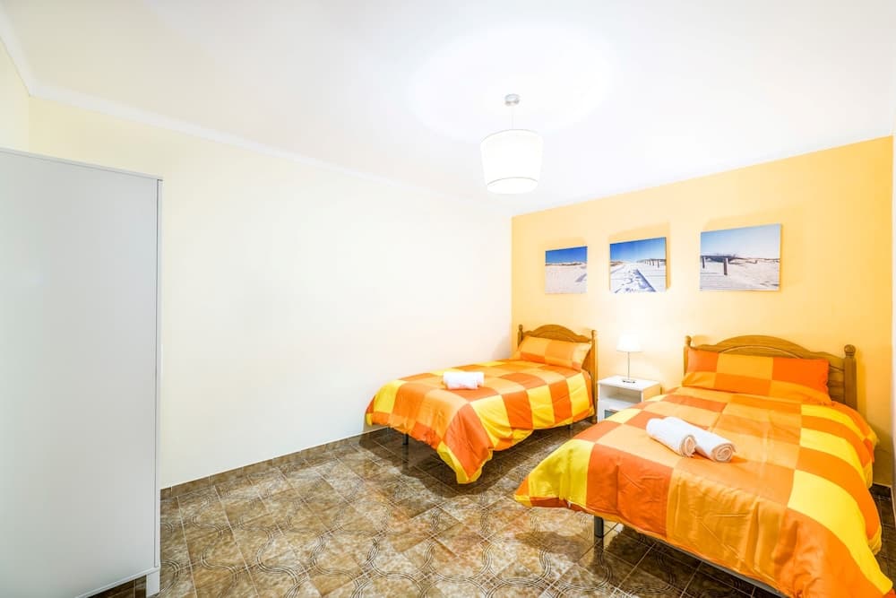 A cozy hotel room features two vibrant orange-and-yellow beds against a warm yellow wall adorned with framed beach scenes, ideal for travelers seeking comfort and a cheerful ambiance.