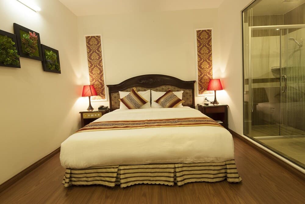 A cozy hotel bedroom features a neatly made bed with red lamps on side tables, framed artwork on the walls, and a glass-enclosed shower, ideal for travelers seeking comfort and elegance.