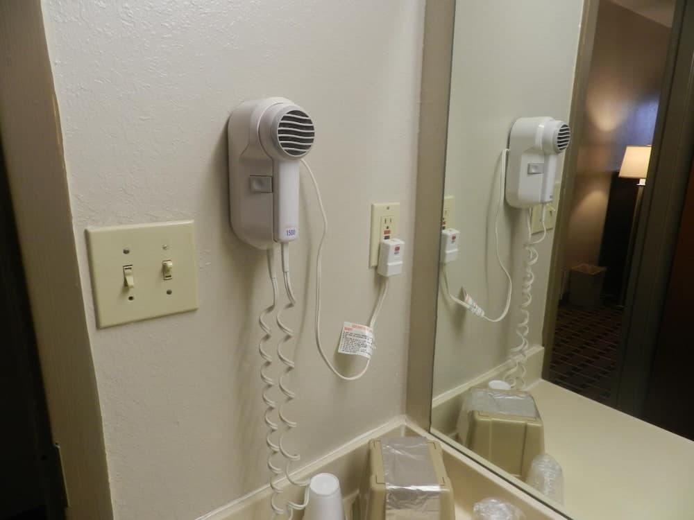 A cozy hotel bathroom corner features a mounted hairdryer, light switches, and a mirror reflecting the hallway, ideal for travelers needing quick grooming amenities.