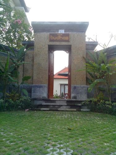 A serene tropical garden features a stone archway labeled "budhas," framing a cozy house through lush greenery, ideal for travelers seeking peaceful accommodation in a natural setting.