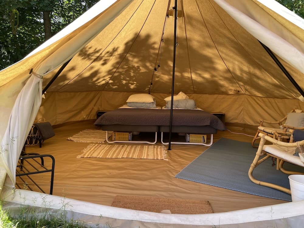 A cozy glamping tent setup with a comfortable bed, rattan chairs, and rugs bathed in dappled sunlight, ideal for a serene outdoor retreat experience.