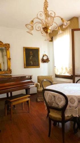 A cozy, vintage living room with warm wooden floors, an ornate chandelier, a grand piano, and lace-covered furniture evokes a nostalgic charm, perfect for travelers seeking a quaint, historical accommodation experience.