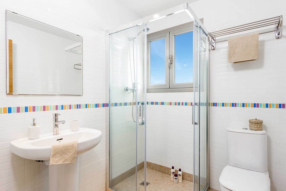 A bright, modern bathroom with white tiled walls, a colorful mosaic border, a glass-enclosed shower, and a sunny window creates a clean and inviting atmosphere ideal for travelers seeking comfort and hygiene.