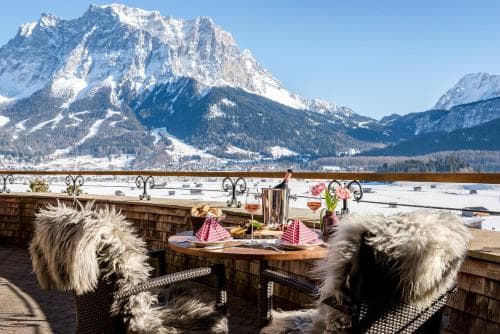 A cozy outdoor dining setup with fur-lined chairs overlooks a stunning snowy mountain landscape, perfect for a luxurious winter getaway experience.