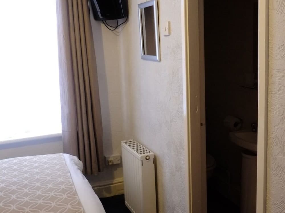A cozy hotel room features a neatly made bed by a bright window, a wall-mounted TV, a radiator, and an adjacent bathroom, ideal for travelers seeking comfort and convenience.