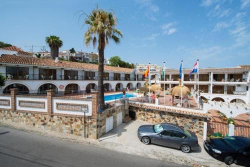 A sunny resort scene featuring a poolside area with palm trees, colorful flags, and parked cars, ideal for travelers seeking a relaxing vacation spot in a Mediterranean-style setting.