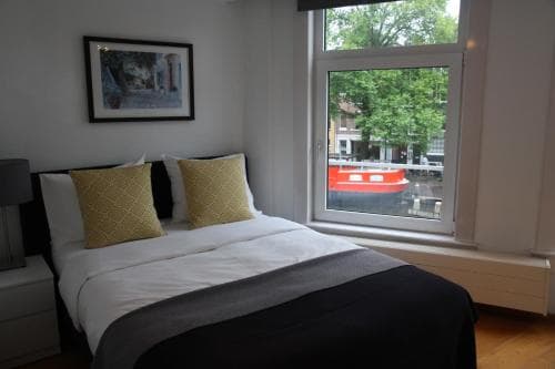 A cozy hotel room with a neatly made bed and yellow pillows overlooks a serene canal scene with a red boat, ideal for travelers seeking comfort and charm in a waterfront setting.