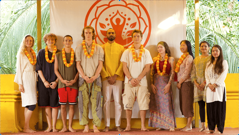 A serene yoga retreat group stands barefoot on a wooden deck adorned with marigold garlands, framed by lush greenery and a vibrant red mandala backdrop, ideal for travelers seeking spiritual growth and wellness experiences in Goa, India.