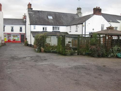 A quaint village courtyard features traditional white buildings with red accents, green trellises adorned with plants, and a paved area, ideal for exploring charming local architecture and enjoying a peaceful retreat.