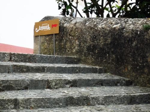 A rustic stone staircase leads up to a textured wall adorned with a wooden sign pointing towards "Mercado Artesanal," evoking a quaint, artisanal market atmosphere ideal for cultural exploration and souvenir shopping.