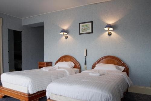A cozy hotel room features two neatly made twin beds with wooden headboards, soft blue walls adorned with framed art and wall-mounted lamps, creating a serene atmosphere ideal for travelers seeking restful accommodations.
