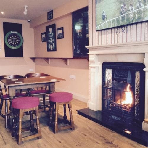 A cozy pub interior features a warm fireplace, wooden tables with pink stools, dartboards on the wall, and a TV showing sports, ideal for travelers seeking a relaxing spot to unwind and socialize.