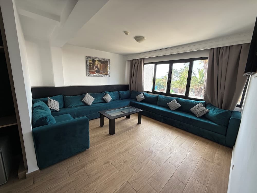 A cozy living room with a plush teal sectional sofa, wooden flooring, and large windows offering a serene view of palm trees, ideal for travelers seeking a relaxing vacation rental.