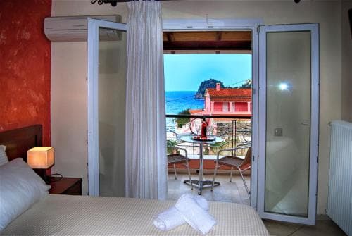 A cozy hotel room with a serene view of the sea and rocky cliffs through glass doors leads to a balcony with chairs, ideal for travelers seeking relaxation and scenic beauty.
