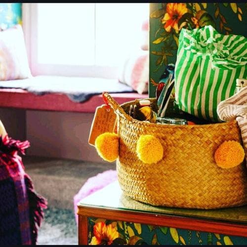 A cozy indoor setting features a vibrant woven basket adorned with yellow pom-poms on a floral table, evoking a cheerful and homey atmosphere ideal for lifestyle photography or interior design inspiration.