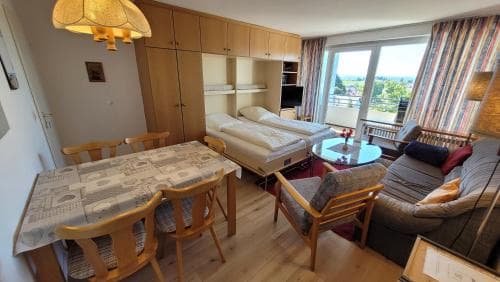 A cozy apartment interior featuring wooden furniture, a dining table, comfortable seating, and large windows offering scenic views, ideal for travelers seeking a homey and relaxing stay.
