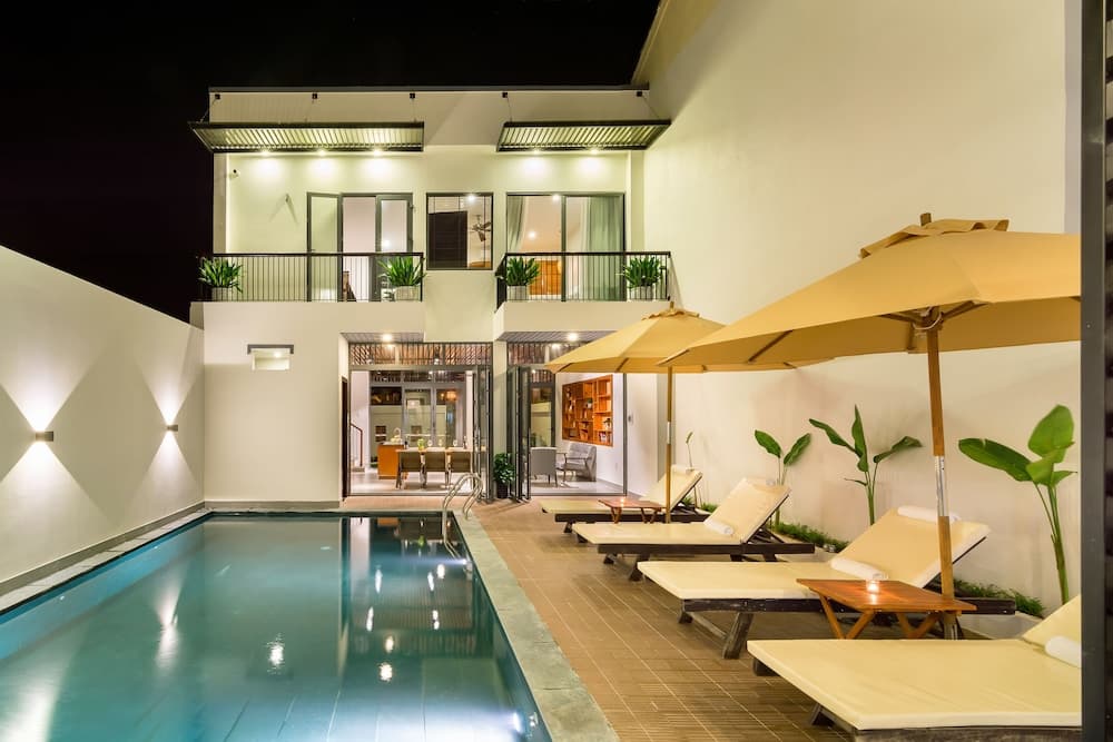 A serene night-time view of a modern villa's private pool area, featuring lounge chairs under umbrellas, lush greenery, and warmly lit interiors, ideal for luxury vacation retreats.