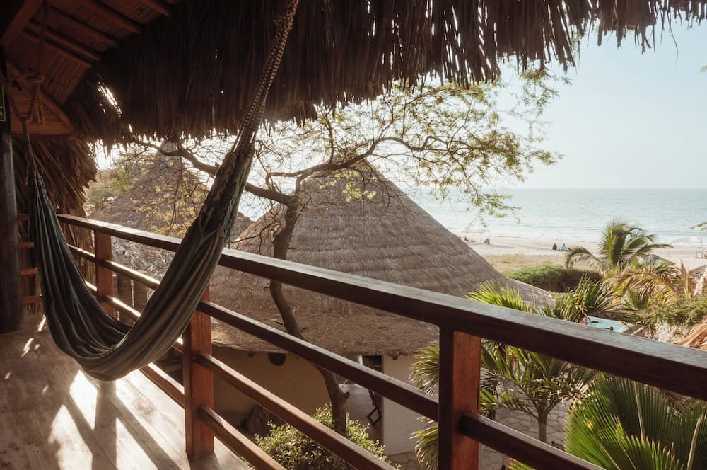 A serene beachside balcony with a hammock overlooks thatched-roof huts and palm trees, offering a tranquil tropical escape ideal for relaxation and vacation photography.