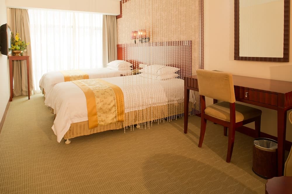 A cozy hotel room features two neatly made beds with yellow accents, a wooden desk with a chair, sheer curtains, and a warm ambiance, ideal for travelers seeking comfort and relaxation.