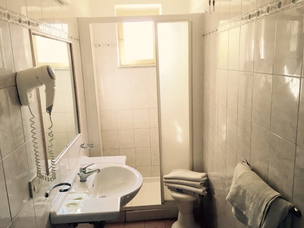 A cozy hotel bathroom features white tiled walls, a sink with a mounted hairdryer, a neatly folded towel on the toilet, and a frosted glass shower door, ideal for travelers seeking cleanliness and convenience.