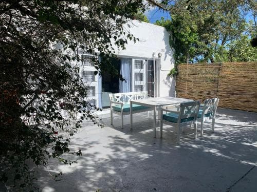 A serene outdoor patio features white furniture under leafy greenery beside a charming white house, ideal for a relaxing vacation retreat.