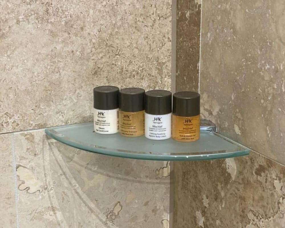 A cozy hotel bathroom corner features a glass shelf holding neatly arranged toiletries against a textured marble wall, ideal for travelers seeking comfort and convenience.