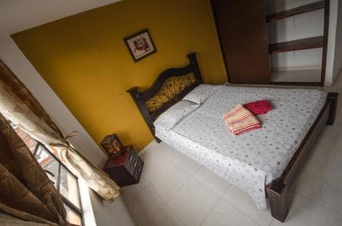 A cozy bedroom with a wooden bed adorned with patterned bedding and vibrant yellow walls, featuring a window with curtains and a small bedside table, ideal for travelers seeking a comfortable and inviting accommodation.