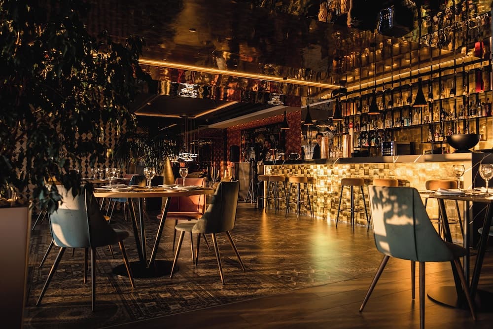 A cozy, warmly lit bar with reflective ceilings, modern furniture, and lush greenery creates an inviting atmosphere perfect for intimate dinners or social gatherings.