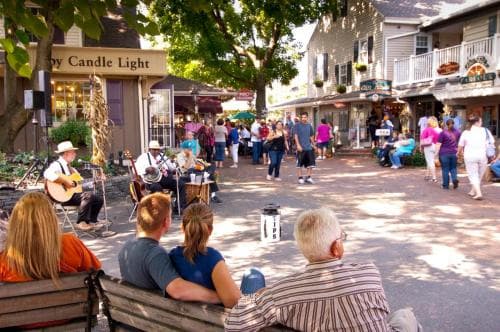 A lively outdoor scene captures a quaint town street lined with charming shops and bustling with visitors enjoying live music from musicians on benches, perfect for a relaxing day trip or exploring local culture.