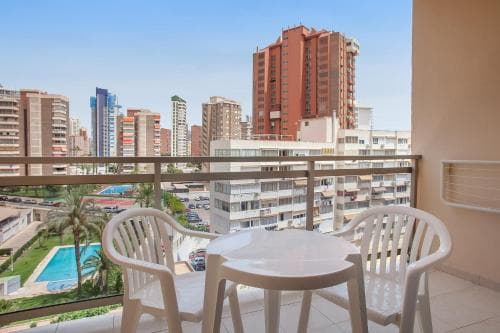 A cozy balcony with white chairs and a table overlooks a vibrant cityscape featuring modern high-rise buildings, a lush green park, and a swimming pool, ideal for travelers seeking urban relaxation and scenic views.