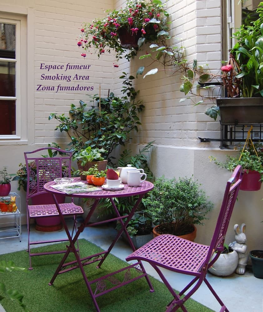 A cozy urban patio features vibrant purple furniture surrounded by lush greenery and hanging flowers, creating an inviting smoking area perfect for travelers seeking a tranquil retreat.
