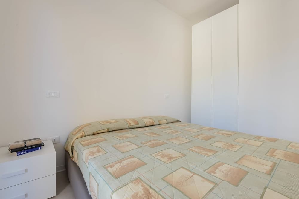 A minimalist bedroom with a neatly made bed featuring a patterned quilt, white furniture including a wardrobe and bedside table with books, exudes a calm and cozy atmosphere ideal for travelers seeking a simple and serene accommodation.