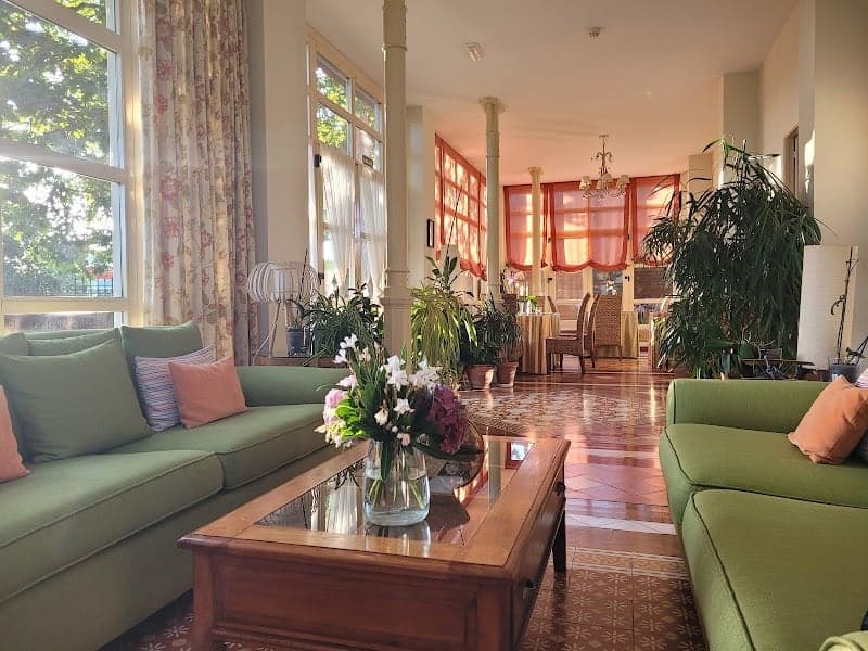 A cozy living room in a charming Spanish casona features vibrant green sofas, lush indoor plants, and large windows with floral curtains, creating an inviting atmosphere perfect for travelers seeking relaxation and comfort.