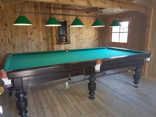 A cozy cabin interior features a classic pool table under green lampshades, ideal for travelers seeking relaxation and entertainment in a rustic setting.