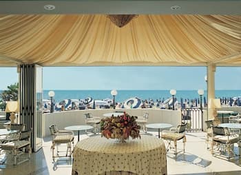 A luxurious seaside café with elegant drapery and outdoor seating overlooks a bustling beachfront, perfect for travelers seeking a relaxing coastal dining experience.