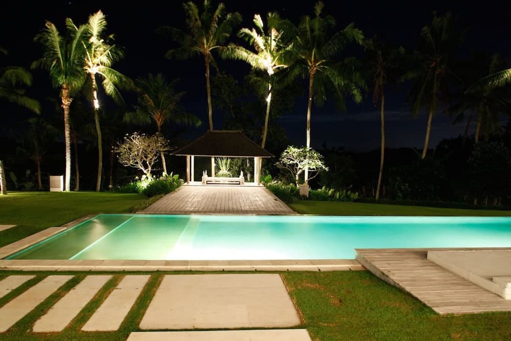 A serene night scene features a glowing pool leading to a tranquil pavilion surrounded by illuminated palm trees, ideal for a luxurious tropical retreat.